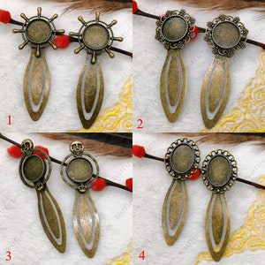 Puede incluir: Cuatro juegos de colgantes de marcapáginas de metal dorado antiguo con diferentes diseños. Cada colgante presenta un cabujón de vidrio redondo u ovalado. Los diseños incluyen una calavera, una rueda y un patrón floral.