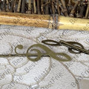 May include: Three antique bronze-colored hair clips. Two clips feature a treble clef design, and the third clip has a similar musical note shape. The clips are on a textured, light-colored surface.