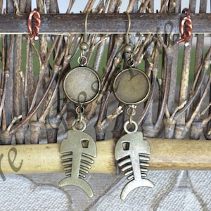 May include: A pair of bronze earrings with a fishbone design and round glass cabochons. The earrings are hanging from a wooden background.