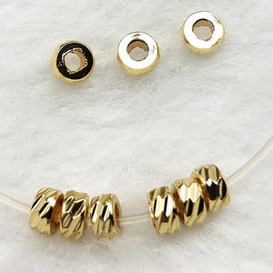 May include: Gold-colored, textured beads with a geometric pattern. The beads are round with a hole through the center, and are strung on a clear cord. The beads are displayed on a white, textured surface.