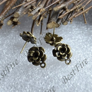 May include: Antique bronze-colored flower-shaped stud earrings. The earrings have a detailed floral design with a post and a small loop at the bottom. The earrings are set against a neutral background with twigs.