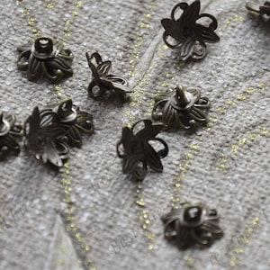 Peut inclure: Dix boutons en métal de couleur bronze antique en forme de fleur avec un motif décoratif. Les boutons ont une boucle sur le dessus pour coudre.