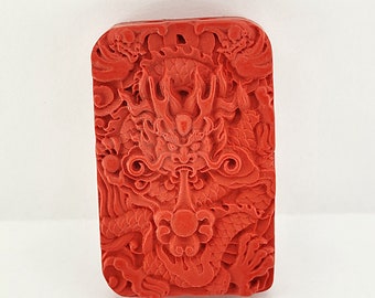 May include: A red carved dragon pendant with intricate details. The dragon's face is prominent, with large eyes and a wide mouth. The pendant is rectangular in shape and has a smooth, polished surface.