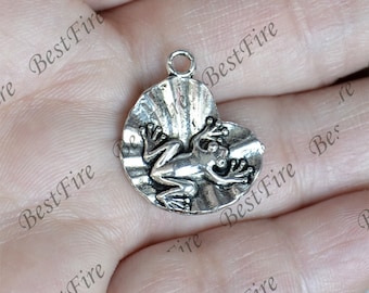 Pode incluir: Encanto de prata com um sapo sentado em uma concha. O sapo é detalhado com pele texturizada e tem um design em relevo nas costas. A concha é texturizada e tem um design em relevo na parte superior.