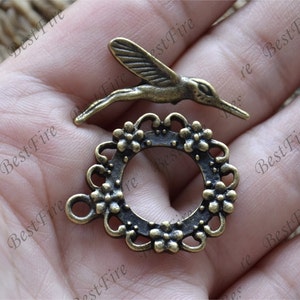 Peut inclure: Deux composants de bijoux en métal de couleur bronze antique. L'un est un colibri, l'autre un cercle floral. Le colibri est en vol, avec des détails sur les ailes et le corps. Le cercle floral a un bord décoratif.