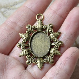 Może przedstawiać: Brązowy wisiorek z ozdobnym wzorem kwiatowym i owalną pustą przestrzenią na kameę lub zdjęcie. Wisiorek ma pętelkę u góry do przymocowania łańcuszka.