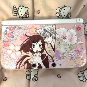 May include: A pink Nintendo 3DS console with an anime design. The artwork shows two characters with floral and leaf details. The console has a white top and a clear protective cover.