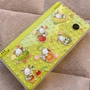 Può includere: Una custodia verde lime per dispositivo di gioco portatile con personaggi di cani dei cartoni animati e frutta. La custodia ha una copertura in plastica trasparente ed è decorata con immagini del cane con fragole, arance e pere. La custodia ha un obiettivo nero.