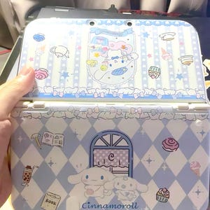 Puede incluir: Una funda de Nintendo 3DS azul claro y blanca con un diseño de Cinnamoroll. La funda está decorada con imágenes de dibujos animados de postres, estrellas y lazos. El texto "Cinnamoroll" está impreso en la parte inferior de la funda.