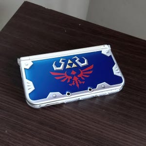 May include: A white and blue Nintendo 3DS handheld game console. The top cover has a blue background with a red and gold emblem. The console is closed and resting on a dark brown surface.