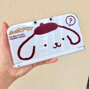 May include: White Nintendo 3DS case with a cartoon character design in maroon and black. The case has Japanese text and the English phrase "Fun days of warm friendship forever!"