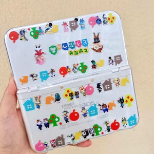 May include: A white Nintendo 3DS LL handheld game console decorated with colorful stickers featuring characters, houses, trees, and fruit. The stickers are from the Animal Crossing series. The console is open and held in a hand.