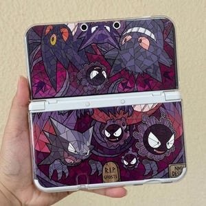 Puede incluir: Una consola de juegos portátil con una cubierta estilo vitral en tonos morados y rosa oscuro. El diseño muestra varias criaturas de tipo fantasma, una lápida con la etiqueta R.I.P. GHOSTS y un pequeño letrero con el texto NMH DESIGNS.
