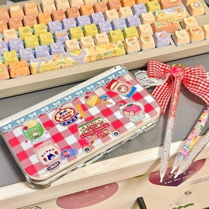 May include: A clear pencil case with a red and white checkered pattern, decorated with food-themed stickers. A keyboard with colorful cat-shaped keycaps is in the background. Two pens with glitter and a red bow are also visible.