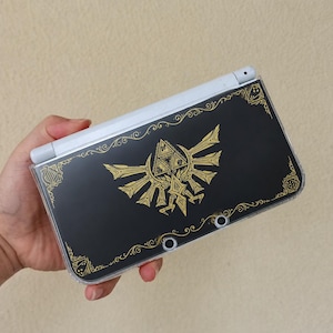 May include: A handheld gaming console featuring a black cover with a gold-coloured Triforce emblem and intricate filigree border designs. The device is encased in a clear protective shell, showing the white hinge and camera lenses on the exterior.