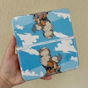 May include: A handheld gaming console featuring a clear protective case with a printed design of the creature Arcanine standing on a rock against a blue sky with white clouds. The design is mirrored on both the top and bottom halves of the device.