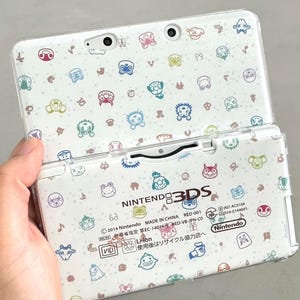 May include: White Nintendo 3DS handheld console with a patterned cover featuring cartoon characters in various colours. The console is open, showing the screen and buttons. The Nintendo logo is visible.