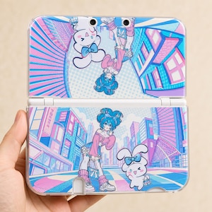 May include: A handheld gaming console featuring a vibrant graphic print of a blue-haired character in a pink sweater and a white rabbit with a blue bow. The design showcases a stylised city background in shades of pink, blue, and white.