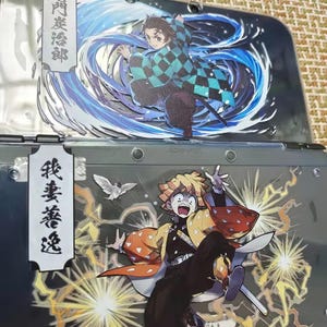 May include: A handheld gaming console with custom decals featuring anime characters. The top screen displays a character in a checkered green and black outfit with blue water effects. The bottom screen shows a character with yellow hair and lightning effects.