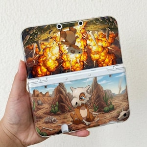 May include: A clear protective case for a handheld gaming console featuring a printed design of a desert landscape with a small brown creature wearing a skull mask. The top section shows a large orange explosion, while the bottom shows the creature.