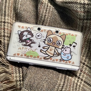 Può includere: Una console di gioco portatile bianca con una cover protettiva trasparente con personaggi di gatti dei cartoni animati e note musicali. Il testo "AIROU FROM THE MONSTER HUNTER" è visibile.