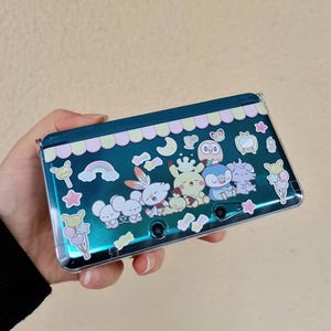 Puede incluir: Una funda teal para Nintendo 3DS con una cubierta de plástico transparente. La funda está decorada con personajes de dibujos animados coloridos, un arcoíris y estrellas. El borde superior presenta un diseño festoneado rosa y blanco.