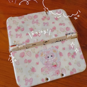 May include: A white handheld gaming console case decorated with pink strawberries and bows. The case has the text "Enjoy!" and a cartoon cat illustration with the text "Strawberry Cats".