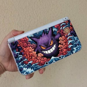 Könnte beinhalten: Eine tragbare Spielkonsole mit einer individuellen Hülle, die ein violettes Gengar-Motiv umgeben von stilisierten roten Wolken und blauen Meereswellen zeigt. Das Gerät hat einen weißen Rahmen, eine transparente Schutzhülle und ein kleines Logo.