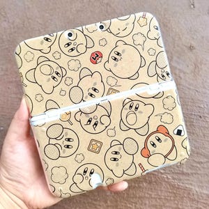 May include: A tan Nintendo 3DS case featuring a repeating pattern of cartoon character illustrations in black outline. The case is open, revealing the interior. The design includes clouds, stars, and food items.