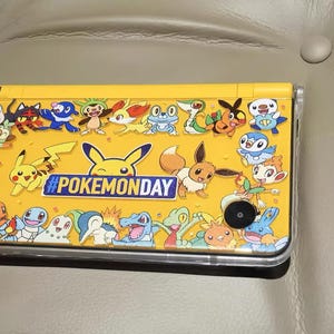 May include: Yellow Nintendo 3DS handheld game console featuring various Pokémon characters and the text "#POKEMONDAY". The console is decorated with images of Pikachu, Eevee, and other popular Pokémon.
