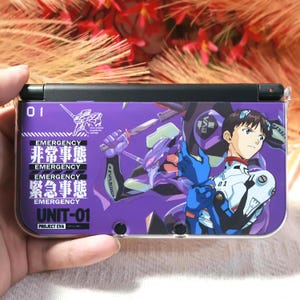Puede incluir: Una funda para consola de juegos portátil morada y negra con personajes de anime y texto japonés. La funda muestra la palabra «EMERGENCY» en inglés y japonés, además de «UNIT-01» y el número «01». El diseño incluye un personaje con un traje azul y blanco.