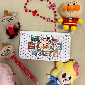 Könnte beinhalten: Eine Sammlung von Anpanman-Artikeln, darunter eine weiße Nintendo DS Lite-Hülle mit roten Punkten und Charaktergrafiken, Plüschtiere, eine rote Perlenkette und ein rot gepunkteter Stift.