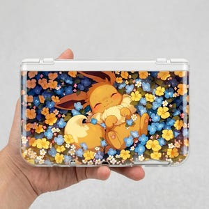 Puede incluir: Una funda protectora transparente para una consola de juegos portátil. La funda presenta un personaje de dibujos animados Eevee acostado en un lecho de flores azules, amarillas y naranjas. La funda está diseñada para un modelo específico de consola portátil.