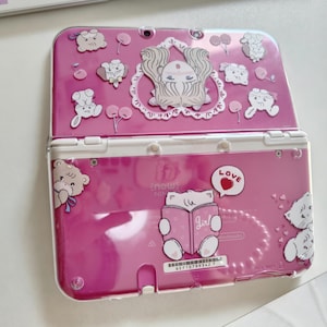 May include: A pink Nintendo 3DS console adorned with stickers of cartoon characters and hearts. The console is open, displaying the screen and buttons. The stickers include the word "Love" in a speech bubble.