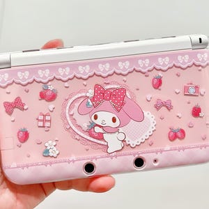 May include: Pink Nintendo 3DS case featuring a My Melody character design with strawberries, bows, and a camera illustration. The case has a clear, protective cover and a white top.