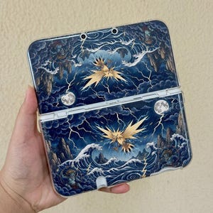 May include: A handheld gaming console case featuring a dark blue graphic print of a golden bird creature amidst stormy clouds, lightning bolts, crashing ocean waves, and full moons. The design includes two small blue figures in the corners.