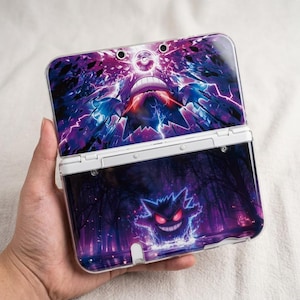 May include: A white Nintendo 3DS handheld game console with a custom cover featuring a purple and blue graphic of a Gengar and other Pokemon characters. The console is open, displaying the screen and buttons.