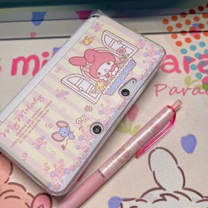 Può includere: Una Nintendo DS Lite rosa e bianca con un design My Melody, con un personaggio dei cartoni animati, fiori e la scritta "My Melody". Una penna rosa con un design abbinato è accanto alla console.