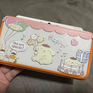 May include: A white and orange handheld gaming device decorated with cartoon characters, balloons, and a birthday cake. The device has the words "You are special!" and "Happy" on it. The device is held in a hand.