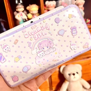 May include: A white and lavender case with a whimsical design featuring a cartoon girl, a teddy bear, and the text "Dreaming Girl." The case is decorated with various cute shapes and polka dots, creating a playful aesthetic.