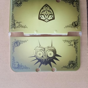 Könnte beinhalten: Eine zweiteilige grüne und transparente Nintendo 3DS-Hülle. Die obere Hälfte zeigt ein schwarzes Wappen und dekorative Eckdesigns. Die untere Hälfte zeigt ein schwarzes Maskendesign. Die Hülle dient zum Schutz des tragbaren Spielgeräts.