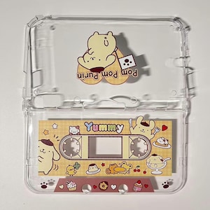May include: A clear plastic cassette tape case with a decorative cassette tape inside. The tape features a cartoon character and the word "Yummy". The case has a cartoon character on the lid. The tape is decorated with food and hearts.