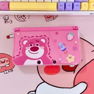 Op de afbeelding: Een roze Nintendo 3DS-hoesje met een cartoon berenontwerp. De hoes heeft een doorzichtige plastic hoes met een aardbei, ster en raket afbeelding. De hoes ligt op een roze ondergrond met andere cartoon afbeeldingen.