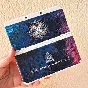 May include: A white and black Nintendo 3DS handheld game console with a custom skin. The skin features a blue and purple gradient design with a gold emblem and the text "MONSTER HUNTER X".