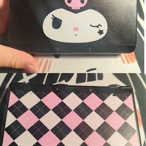 May include: Black Nintendo 3DS console with a Kuromi character design on the top. The design features a pink skull and a white face with a winking eye. The bottom of the console has a pink, black, and white argyle pattern.