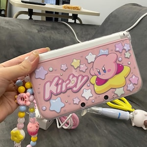 May include: Pink handheld gaming console case with a Kirby design, stars, and the word "Kirby". The case has a white border. A beaded charm bracelet with colourful beads is attached. The console is held in a hand.