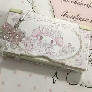 Może przedstawiać: Jasnoróżowe etui na Nintendo DS z postacią z kreskówki, kwiatowym wzorem i napisem "Happy Birthday". Etui ma przezroczystą plastikową osłonę i różowy urok kokardy.