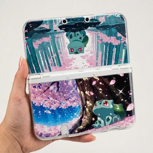 May include: White Nintendo 3DS handheld game console with a custom cover featuring a Bulbasaur character in a cherry blossom forest scene. The top screen shows an upside-down Bulbasaur. The bottom screen shows Bulbasaur sitting under a tree.