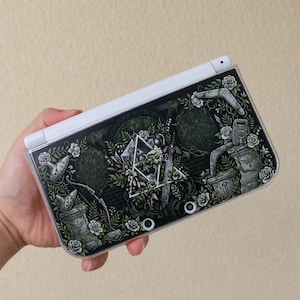 Peut inclure: Une console de jeu portable blanche dotée d'un habillage décoratif avec une illustration botanique vert foncé et noire. Le motif comprend des roses blanches, une épée, un boomerang, une ocarina et un emblème triangulaire central entouré de feuillage.
