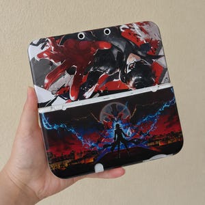 May include: A handheld gaming console case featuring a vibrant anime-style design. The top half displays a character with red and black accents, while the bottom half showcases a silhouette against a cityscape with lightning.